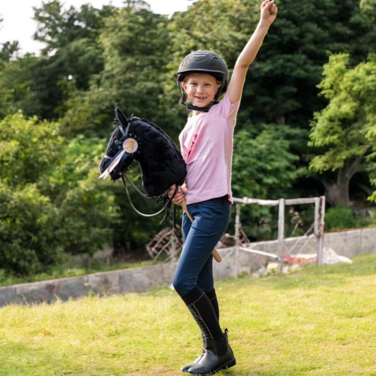 Turnierschleife -Hobby Horsing-, keine Farbe, Kind mit Hobby Horse und erhobenem Arm im Freien.